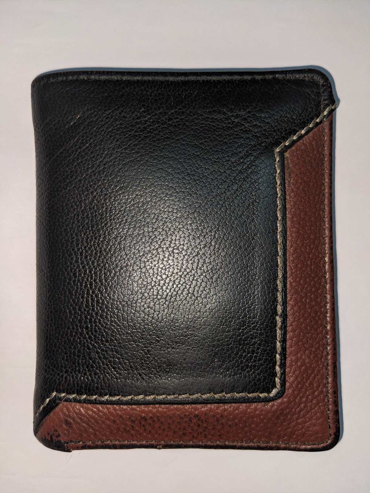 A black and brown bifold leather wallet seen from the front, with visible grain and pale contrast stitching along the panel edge.