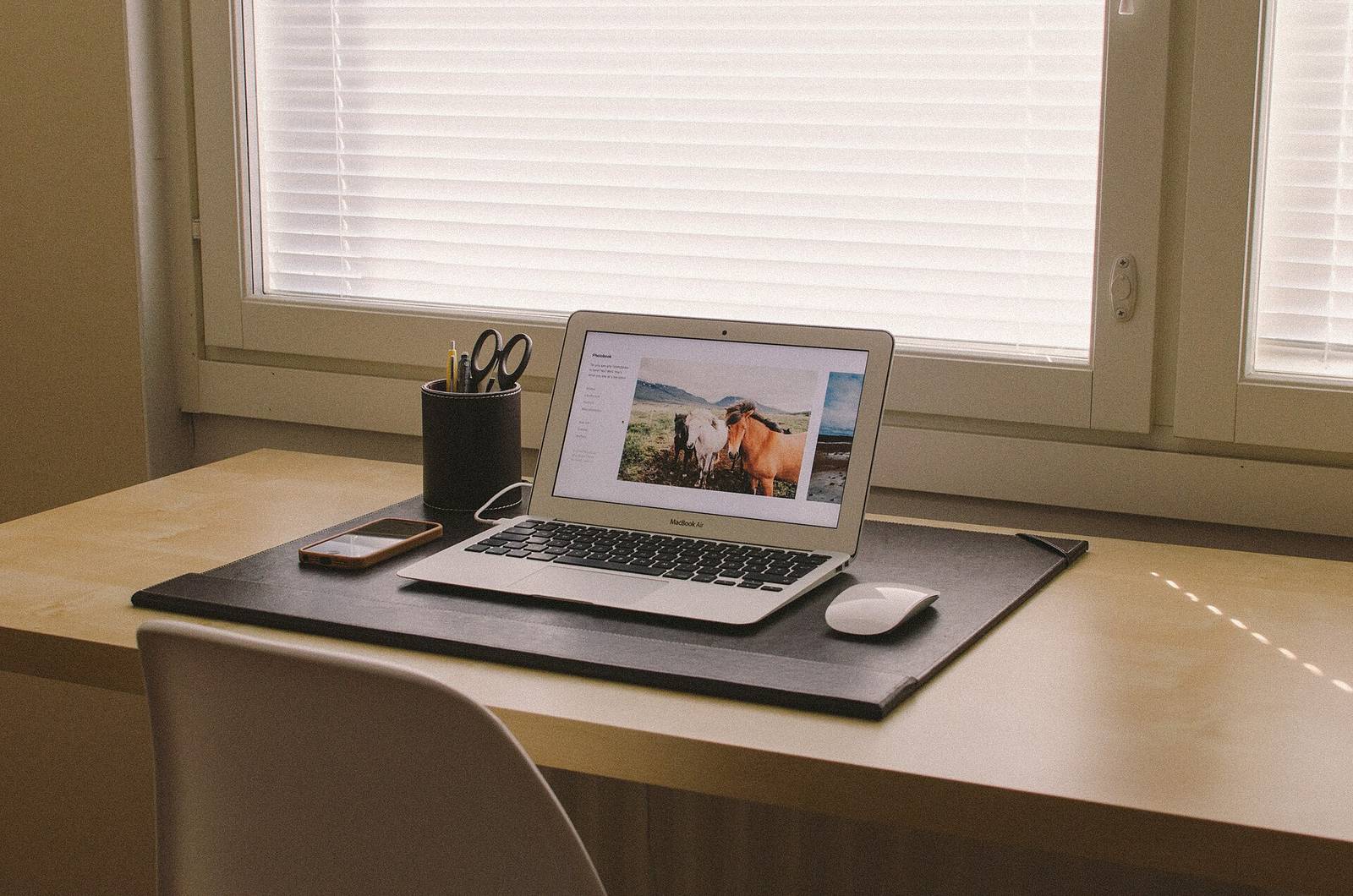 A laptop on a dark desk pad by a window, with a smartphone in a tan case lying beside it and a leather pen cup behind.